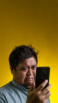 A repulsed Asian male stares at a smartphone held in one hand against a bright yellow background, displaying a strong facial expression of intense disgust and rejection. The individual
