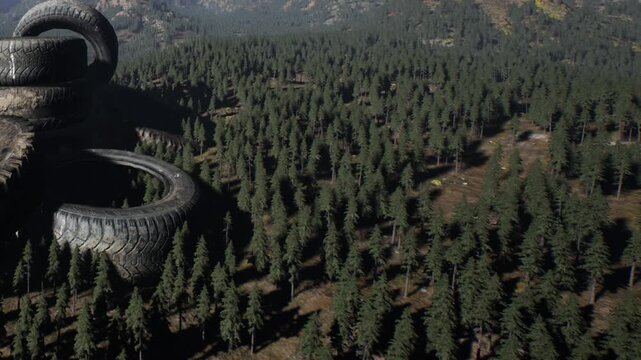 A group of stacked tires sits on the edge of a forest. The area is filled with numerous trees and distant mountains can be seen in the background, suggesting a remote location.