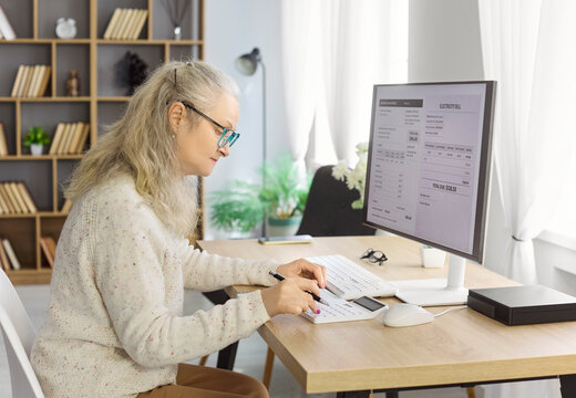 Senior woman reviews bills with calculator. At a home desk she checks an electricity utility bill, and plans monthly expenses to manage finance. Clear concept of home budget and payment.