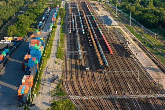 Poland - 14 July 2026: Aerial view of a busy railway freight yard with numerous cargo trains, colorful shipping containers stacked in rows, and surrounding green trees under a clear sky.