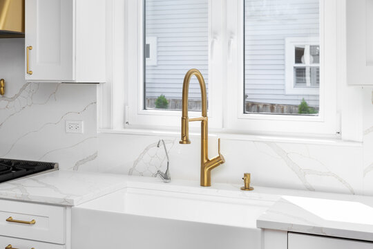 A gold kitchen faucet detail with a marble slab countertop and backsplash and a white farmhouse sink.