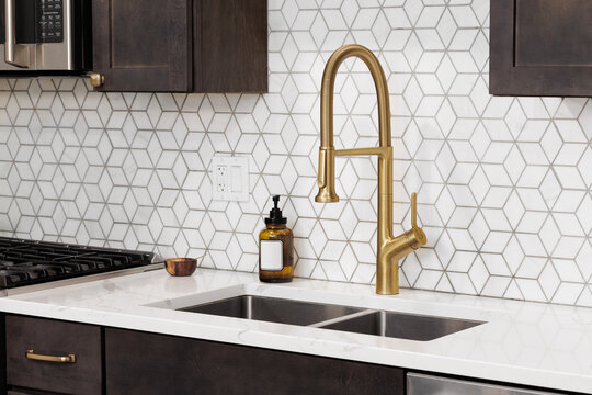 A gold kitchen faucet detail with a mosaic marble tile backsplash, white marble countertop, and dark wood cabinets.