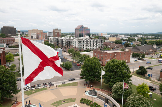 Huntsville, Alabama, United States - 03 June 2022: Aerial view of the Alabama state flag flying over the downtown cityscape with urban buildings, streets, and trees under a cloudy sky.