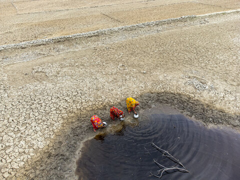 Shyamnagar, Khulna Division, Bangladesh - 25 April 2025: Aerial view of three women in colorful saris collecting water from a small, muddy pool in a dry, cracked landscape.