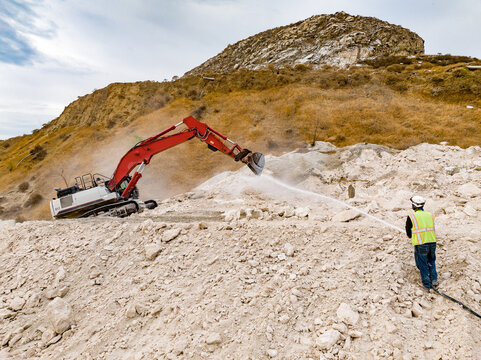 Excavator Breaking Rock While Worker Sprays Water for Dust Suppression