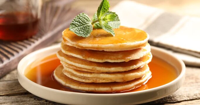 Man adding mint to pancakes with maple syrup at wooden table, closeup