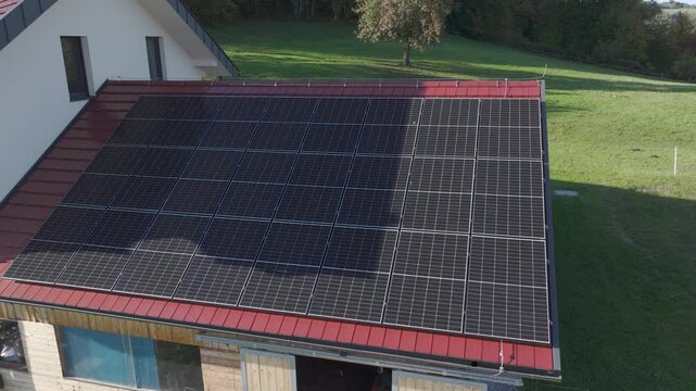 AERIAL: Large rooftop solar panel installation on a modern countryside house. Efficient photovoltaic array covers most of the roof, surrounded by green grass and rural landscape on a bright sunny day.