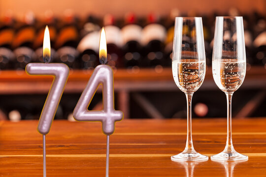 Birthday card, anniversary or celebration of a special day, lit candles forming the number 74 and two champagne glasses; Photo on a background of a bookshelf with blurred wine bottles.