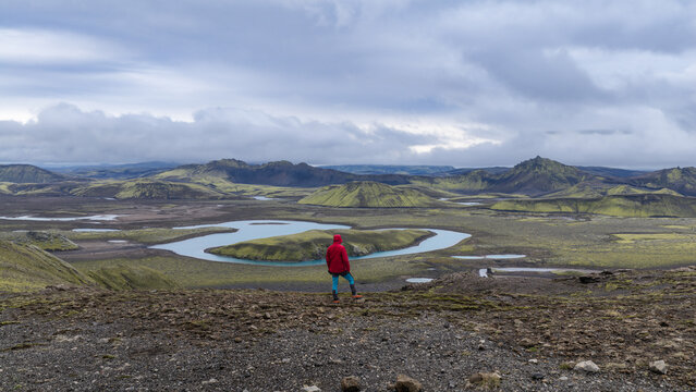 Aerial view of a person in a red jacket standing on a rocky ridge overlooking the winding Skafta river, surrounded by moss-covered volcanic hills and a vast landscape in Skaftarhreppur, Iceland.