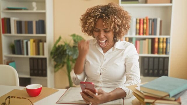Young african american woman smiling, fist pump and hand on chest while reading smartphone with open notebook and stacked books at a building desk; joy achievement.