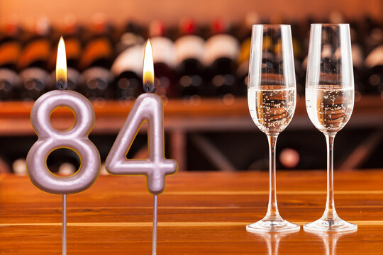 Birthday card, anniversary or celebration of a special day, lit candles forming the number 84 and two champagne glasses; Photo on a background of a bookshelf with blurred wine bottles.