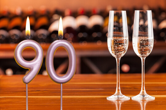 Birthday card, anniversary or celebration of a special day, lit candles forming the number 90 and two champagne glasses; Photo on a background of a bookshelf with blurred wine bottles.