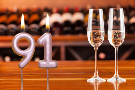 Birthday card, anniversary or celebration of a special day, lit candles forming the number 91 and two champagne glasses; Photo on a background of a bookshelf with blurred wine bottles.