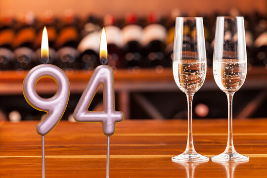 Birthday card, anniversary or celebration of a special day, lit candles forming the number 94 and two champagne glasses; Photo on a background of a bookshelf with blurred wine bottles.
