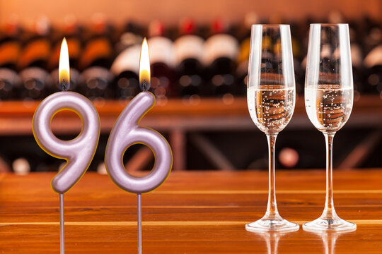 Birthday card, anniversary or celebration of a special day, lit candles forming the number 96 and two champagne glasses; Photo on a background of a bookshelf with blurred wine bottles.