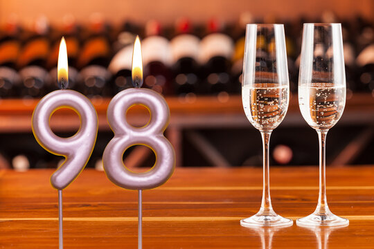 Birthday card, anniversary or celebration of a special day, lit candles forming the number 98 and two champagne glasses; Photo on a background of a bookshelf with blurred wine bottles.