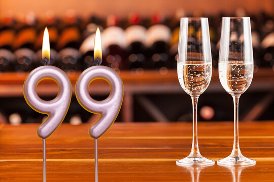Birthday card, anniversary or celebration of a special day, lit candles forming the number 99 and two champagne glasses; Photo on a background of a bookshelf with blurred wine bottles.