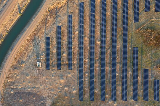 Aerial view of solar panels arranged in long rows at the Boulder Reservoir, with a nearby irrigation canal and dry terrain in Boulder, Colorado, United States.