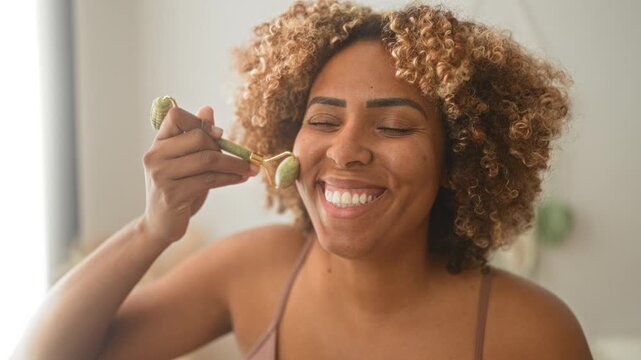 Woman rolling a jade roller across her face in a bright bedroom, smiling with closed eyes and curly hair visible while performing a morning skincare ritual; skincare joy.