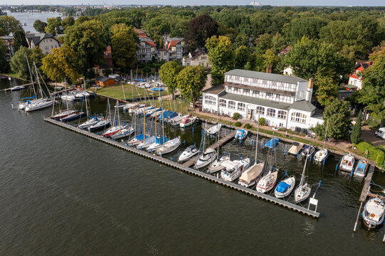 Aerial view of Yachtclub Wendenschloss e.V. with numerous sailboats moored at the pier, surrounded by lush trees and residential buildings along the riverbank in Berlin, Berlin, Germany.