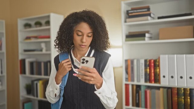 Woman in school uniform holds smartphone and grips backpack strap while reading screen in a building lined with bookshelves; focused study.