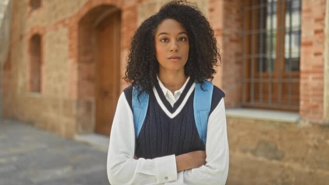 Woman in a school uniform with blue backpack and arms crossed standing before a brick building, gazing to the side with a thoughtful expression; contemplation.