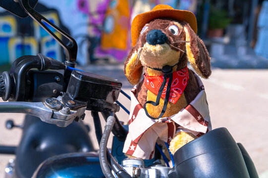 A plush dachshund dog dressed as a cowboy with felt hat, red bandana and star print poncho perches near the handlebar of a parked motorbike, a whimsical mascot for a road culture brand.