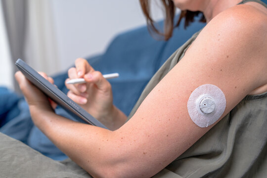 A woman with a continuous glucose monitor on her arm uses a stylus on a digital tablet while seated on a sofa, a concept of diabetes management and health technology.