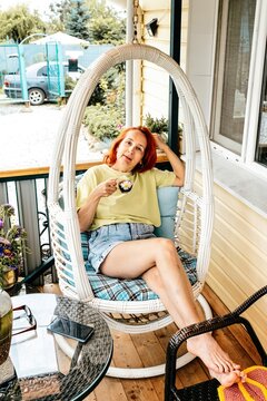 Cozy portrait of a 50 years old russian woman drinking coffee in a hanging chair on a veranda during a summer vacation in Russia