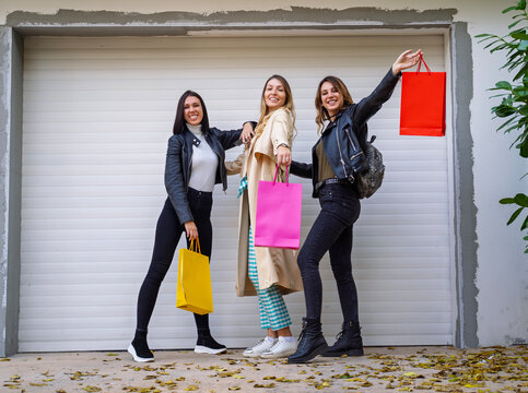 Three smiling young women pose together outdoors while playfully holding up colorful shopping bags, Autumn fashion shopping concept 