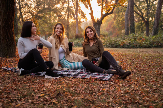 Three young women smile and enjoy warm drinks while relaxing on a picnic blanket in an autumn park filled with fallen leaves