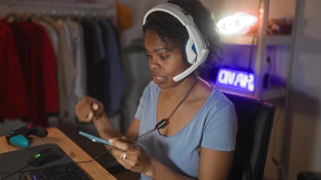 Young woman gamer wearing headset holds smartphone and gamepad, fingers tapping screen at a desk in studio with neon on air sign; concentration.