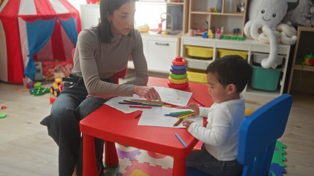Woman guiding young boy drawing with pencil at a red playroom table surrounded by toys, shelving and a stacking ring toy; nurturing calm.