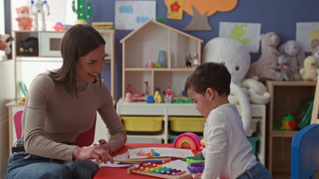 Woman handing xylophone mallet to little boy at table in a playroom surrounded by toys and drawings; early learning joy.