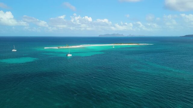 Aerial parallax shot shows sailing boats approaching the narrow white sandbar of Sandy Island off Carriacou, Grenada surrounded by turquoise Caribbean water