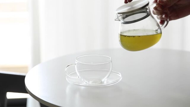 Hand pouring green tea from a clear glass teapot into a glass cup until full on a white table