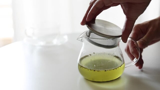 Hand removing the strainer lid from a glass teapot filled with green tea on a white table