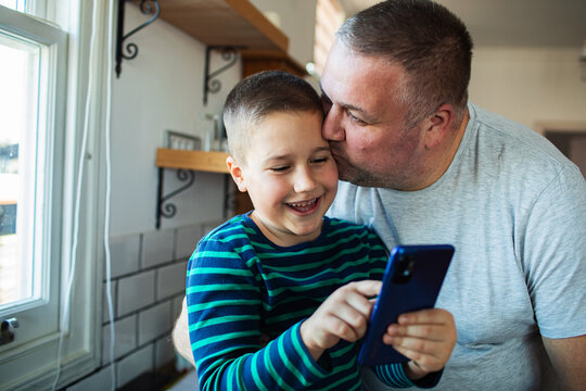 Affectionate father kissing son while using smartphone in kitchen