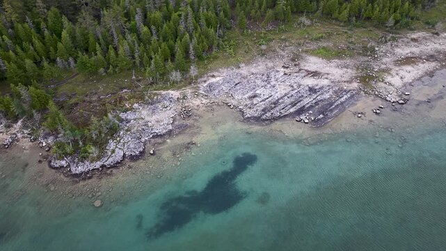 Top down aerial reveals turquoise Lake Huron water dark underwater patches and a rugged forested rock shoreline in Michigan.