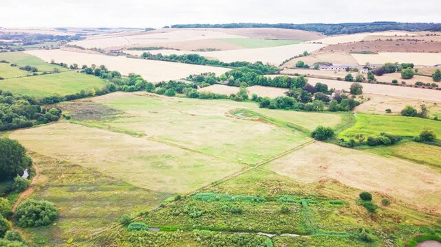 Wide aerial drone shot of rolling chalk downland countryside, showing a patchwork of golden stubble fields and green pasture, hedgerows, a farmstead, and a faint circular crop mark in one field.