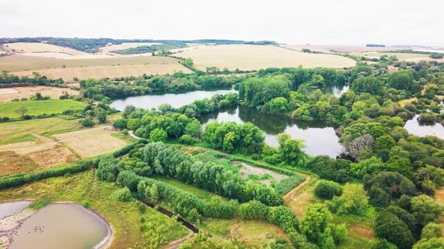Wide drone footage of a chain of wooded lakes bordered by hedgerows, a small paddock, and farmland, with rolling chalk downland fields in the distance.