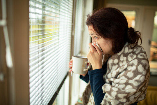 Sick woman with tissue and tea by home window