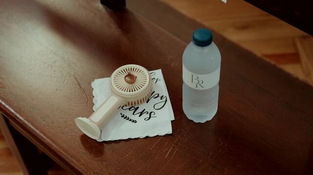 Handheld fan on a printed tissue beside a monogrammed water bottle on a wooden pew