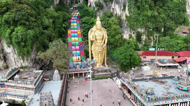 Aerial side view showing drone panning past the giant golden Lord Murugan statue and colorful rainbow stairs at Batu Caves in Malaysia