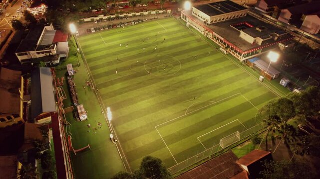 Aerial view of an illuminated soccer field with multiple players participating in football activities after dark, showcasing urban recreation, team sports and nighttime training.