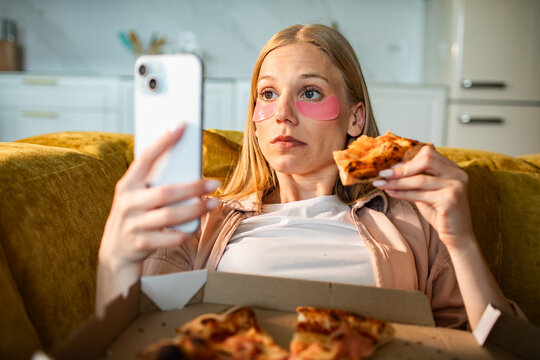 Young woman relaxing on sofa at home with eye patches, eating pizza and using smartphone