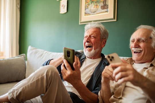 Two senior men laughing with smartphones on home sofa