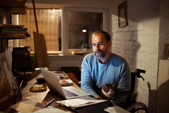 Middle aged man in wheelchair working remotely at home office with cat at night