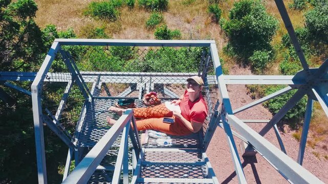 A backpacker resting on a metal staircase of a watchtower and smiling at camera with a wild nature backdrop.