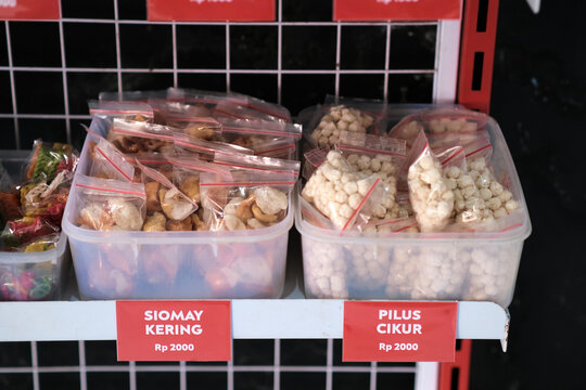 Close-up of plastic ziplock bags containing dried siomay and pilus cikur snacks organized in clear storage tubs with red price labels on a wire rack
